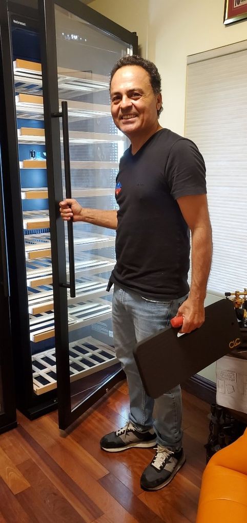 A smiling person stands in an indoor room, holding a black tray while opening the glass door of a tall, multi-shelf cooler.