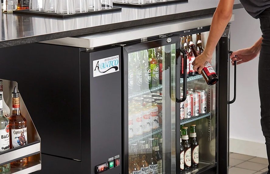 A person reaches into a black under-counter glass-door bar refrigerator to retrieve a bottle.