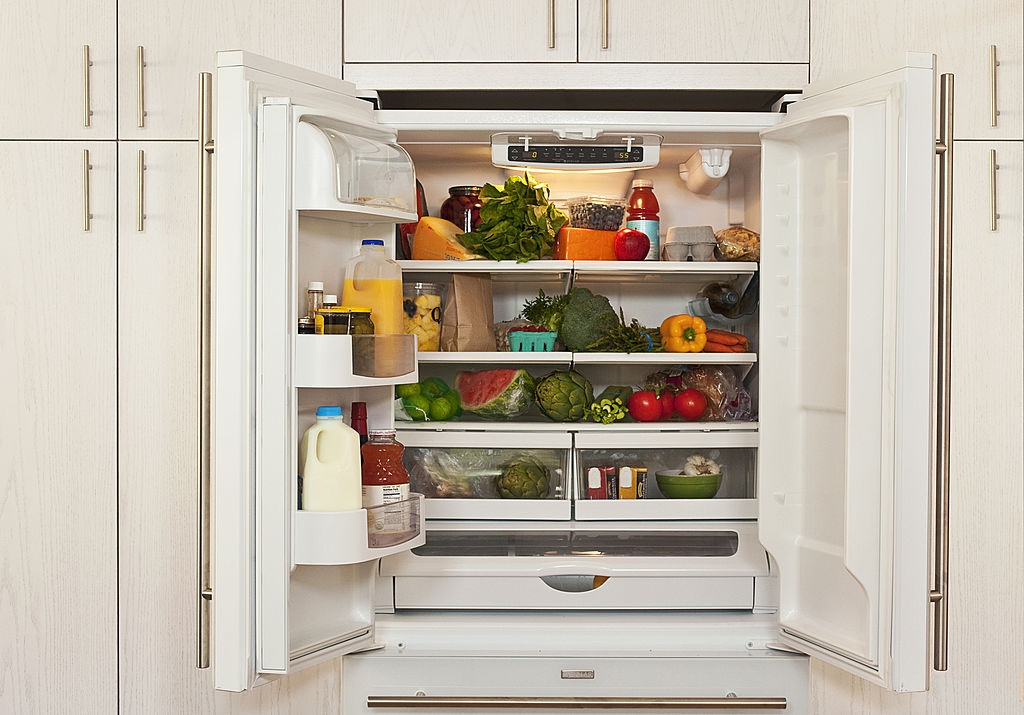 An open refrigerator filled with fresh produce, groceries, and drinks in a modern kitchen with white cabinetry.