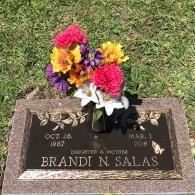 A flat bronze grave marker for Brandi N. Salas (1987–2018) with a bouquet of colorful artificial flowers in a vase.