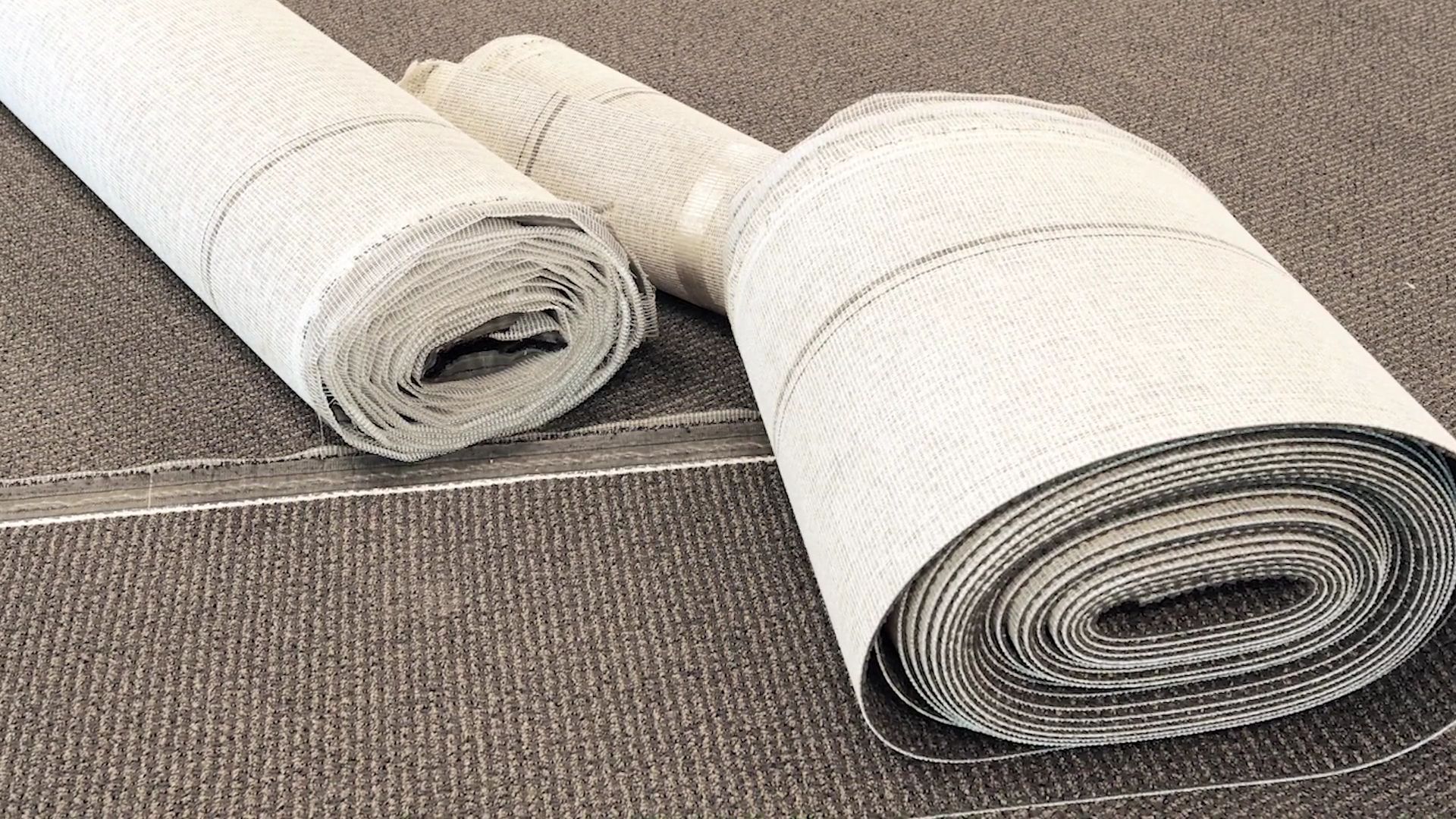 Rolled white carpet samples on a gray carpeted floor, one large roll in the foreground and one smaller roll behind it