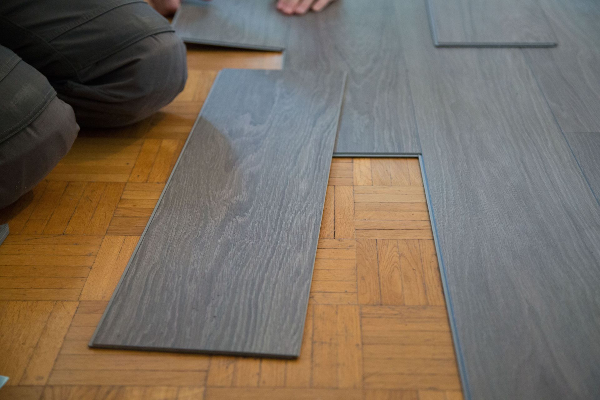 Two people laying gray wood-look flooring over a parquet floor, with one plank partially lifted