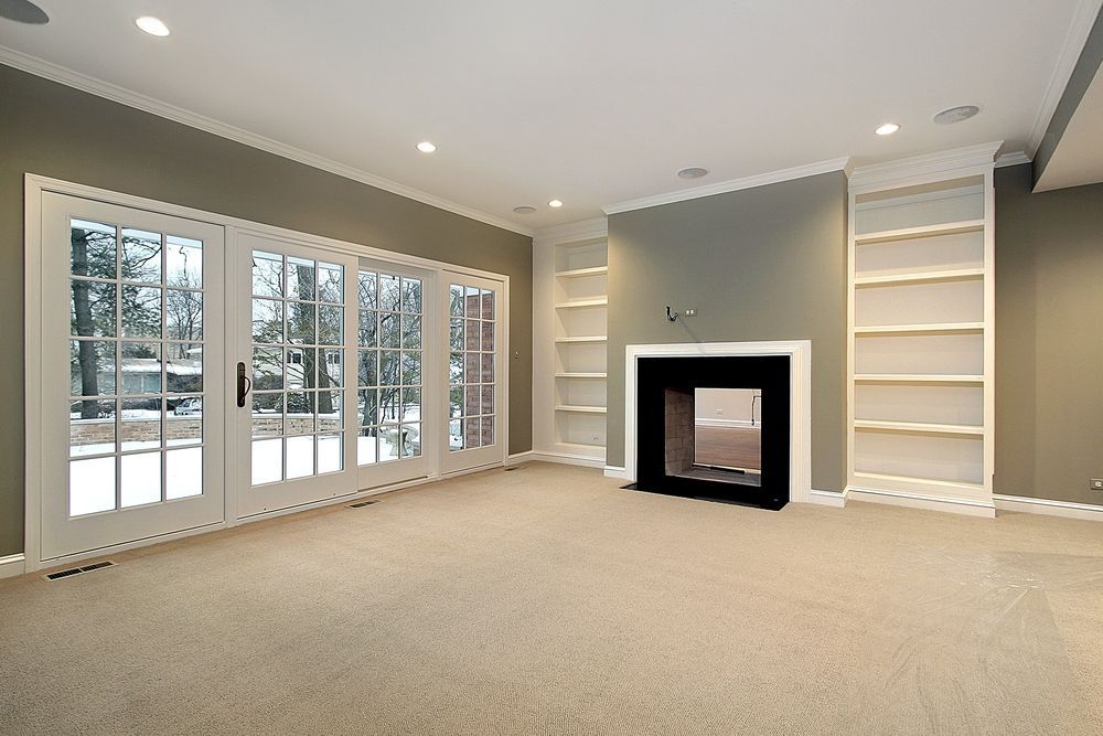 Spacious empty living room with beige carpet, fireplace, built-in shelves, and French doors to snowy outdoors