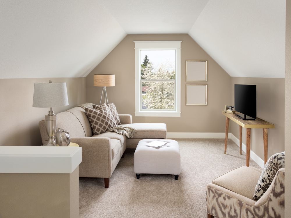 Cozy attic living room with beige sofa, ottoman, TV stand, lamps, and a window.