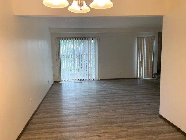 Empty living room with wood-look flooring, beige walls, ceiling lights, and sliding glass doors with vertical blinds.