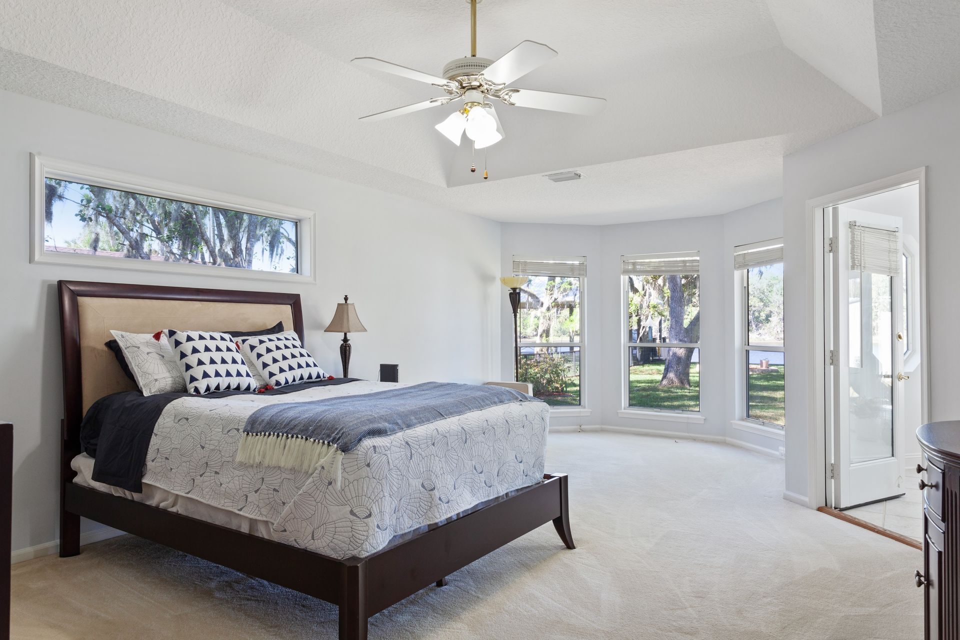 Bright bedroom with a dark wood bed, white walls, ceiling fan, and large windows overlooking a yard