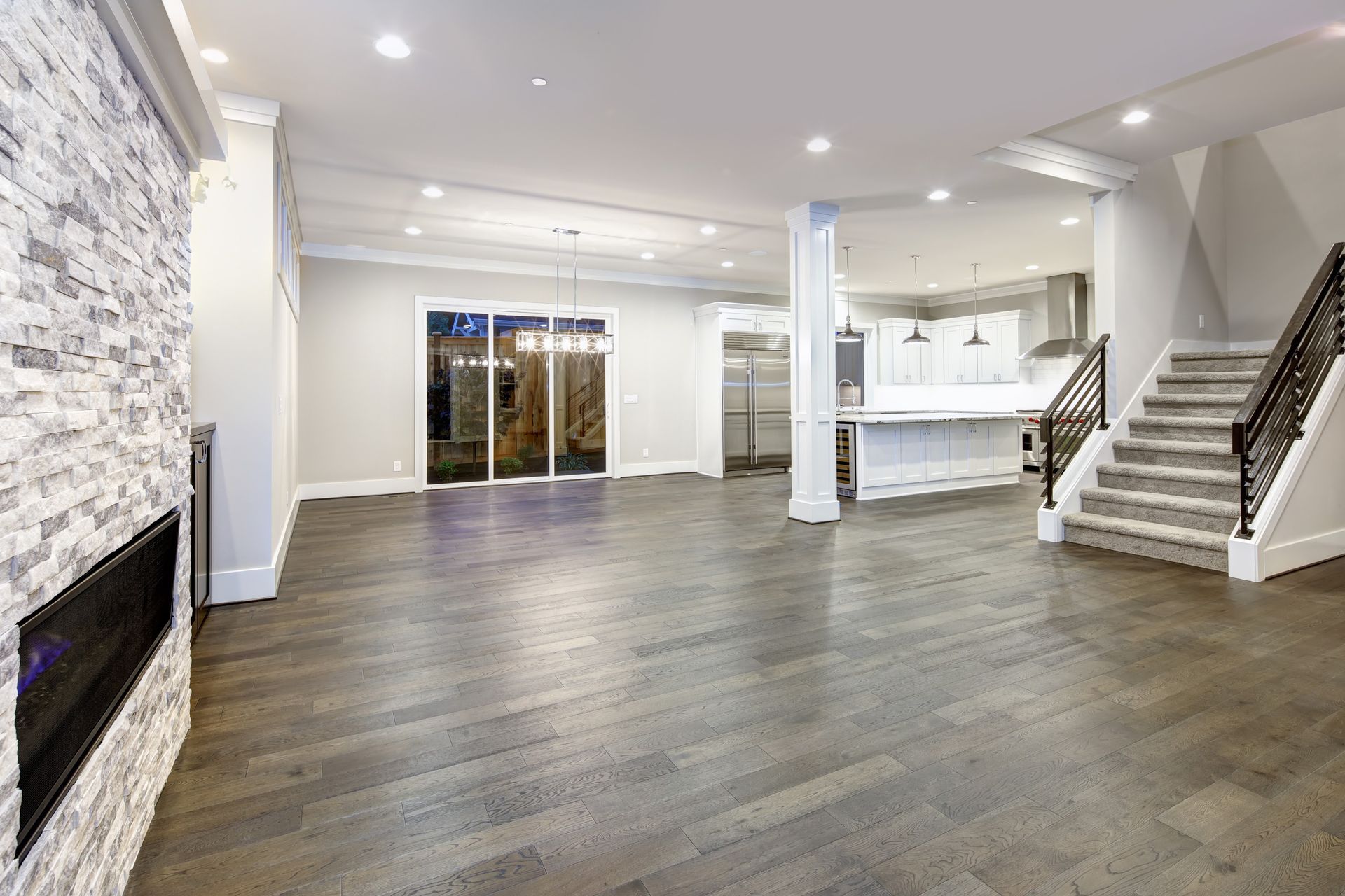 Spacious modern living room with stone fireplace, hardwood floors, white walls, and staircase to the right