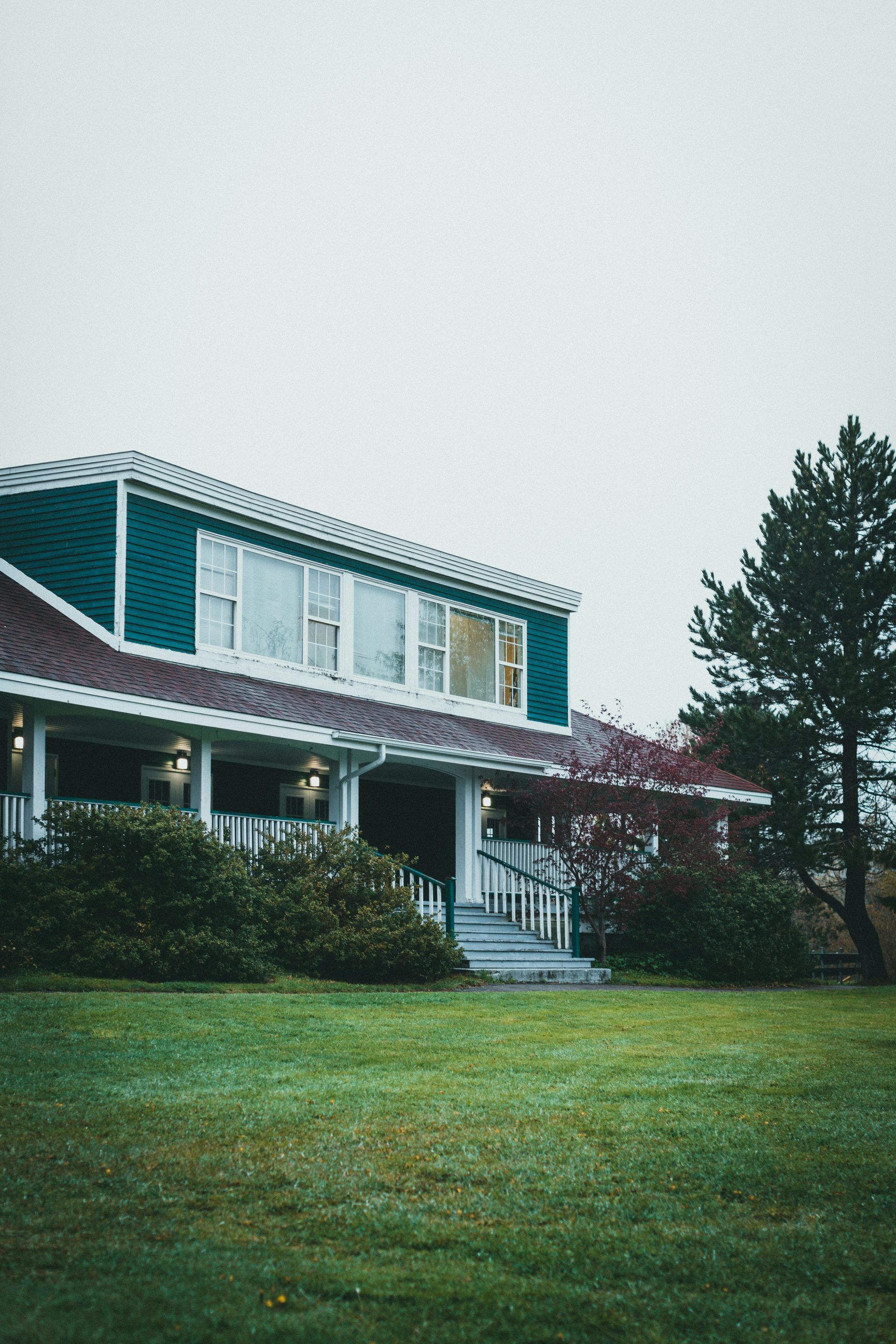 Suburban house with a green lawn, white porch, and tall trees under an overcast sky