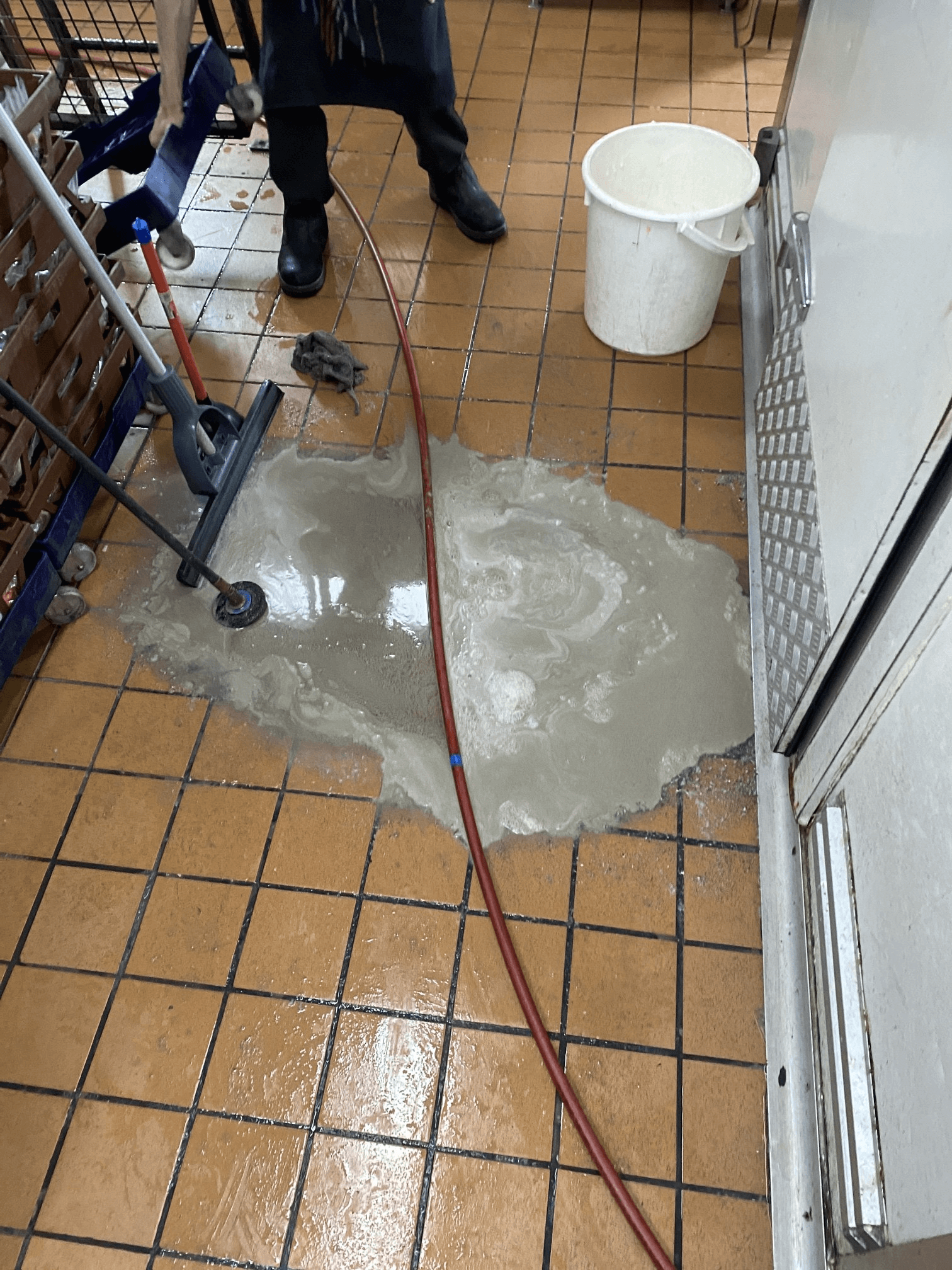 A person uses a floor squeegee to move a large puddle of water across a tiled floor in a commercial kitchen area.