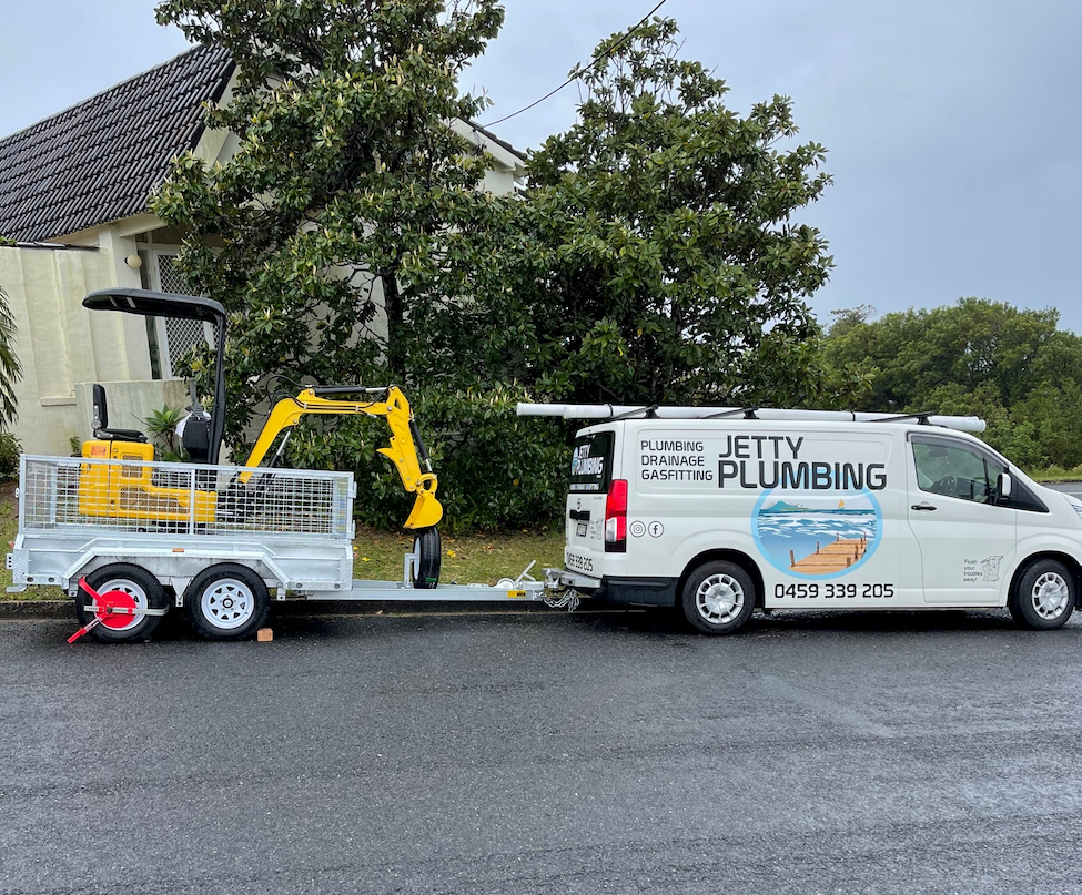 A plumbing van with a trailer carrying a small excavator parked on a street.