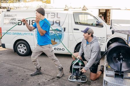 Two technicians in work clothes operate plumbing equipment next to a service van with branding on the side.