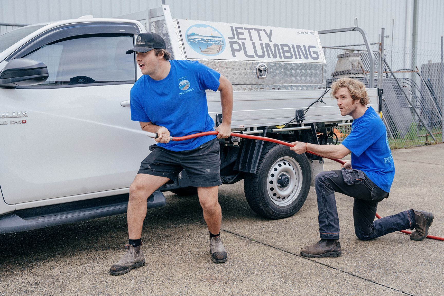 Two people in blue shirts are pulling a red hose connected to a Jetty Plumbing utility truck parked outdoors.