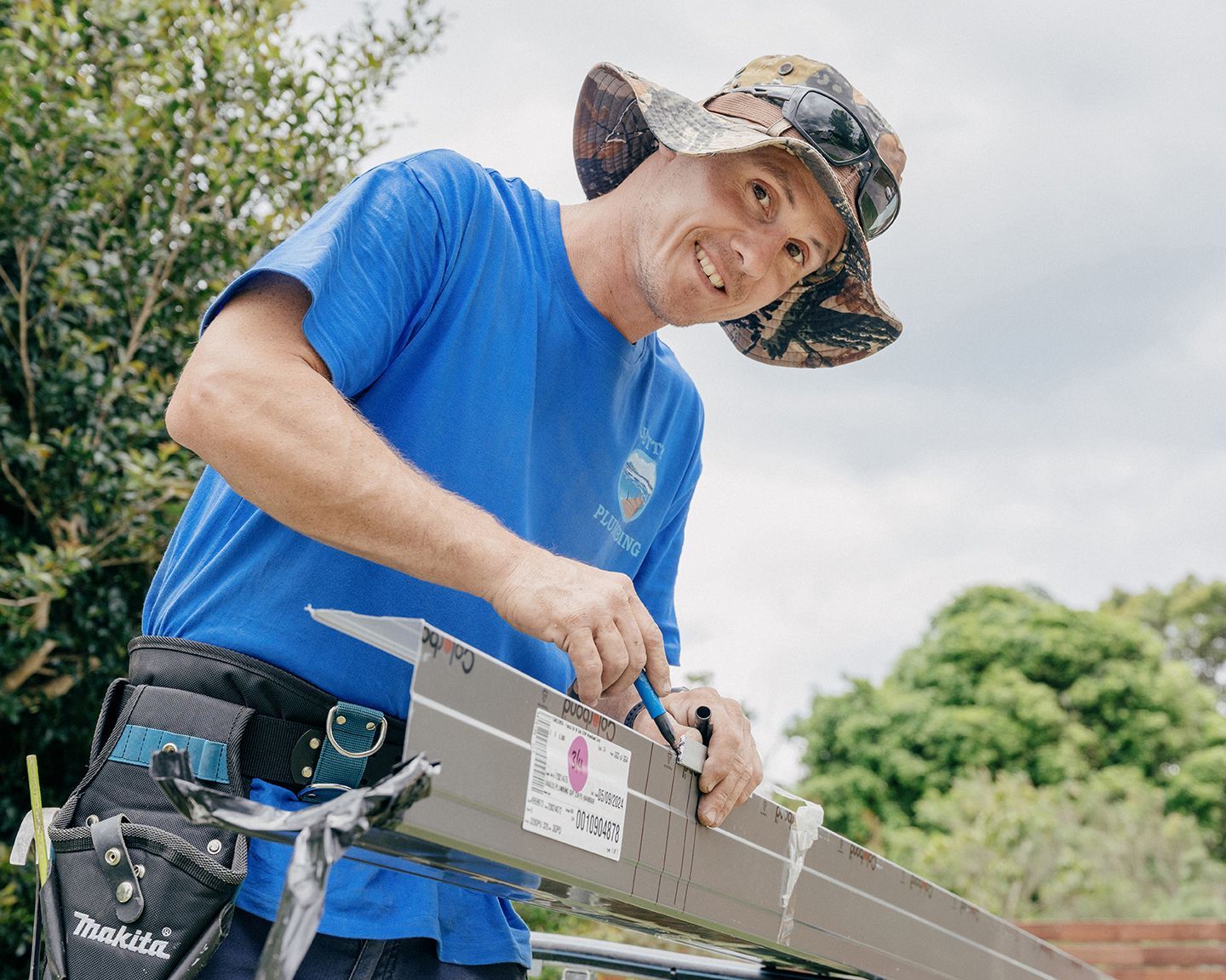 A Jetting plumbing plumber wearing a blue shirt and sun hat works on a piece of metal guttering outdoors, looking at the camera.