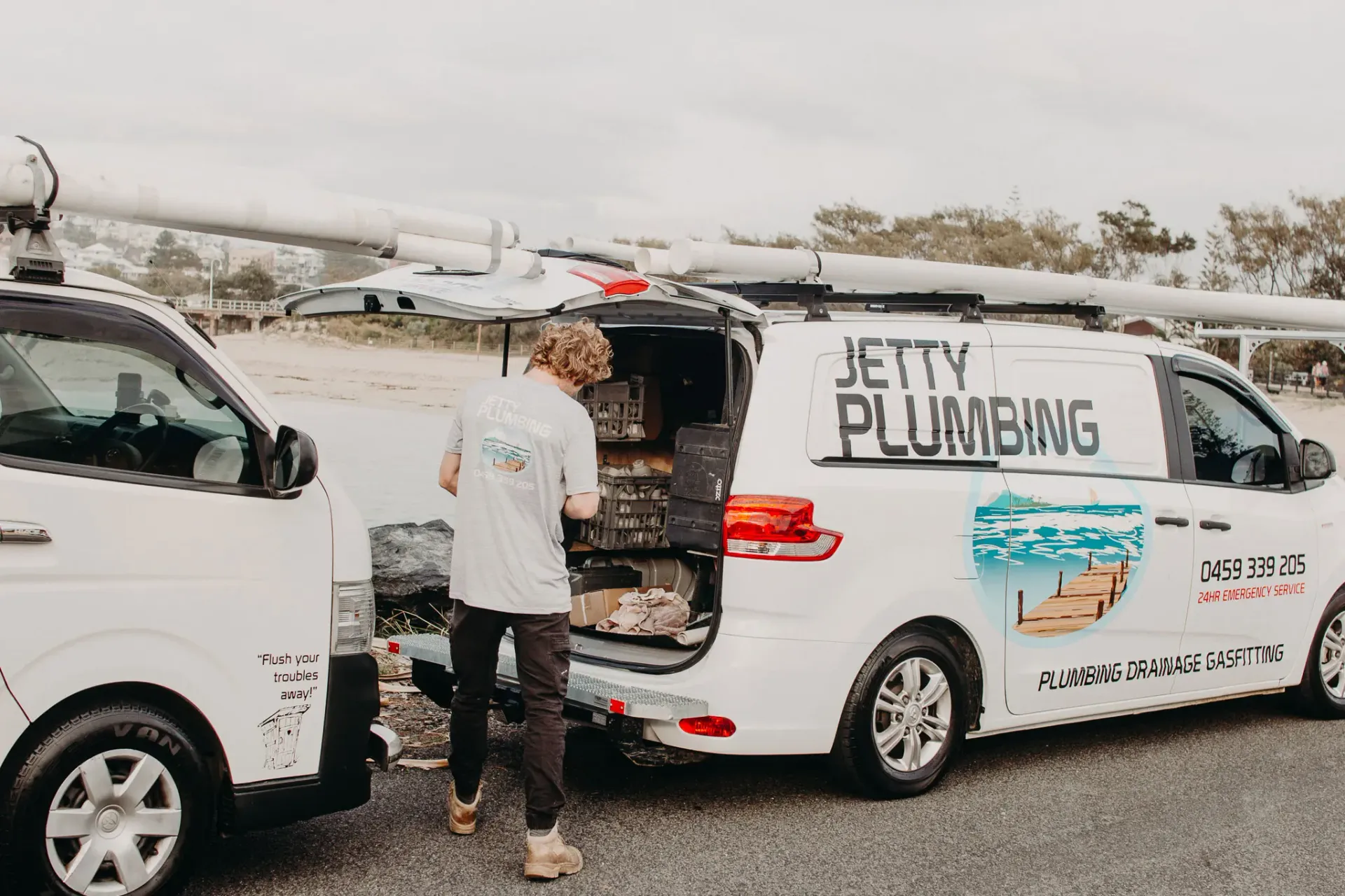 A person stands at the open back of a white Jetty Plumbing van with roof racks parked on a gravel surface.