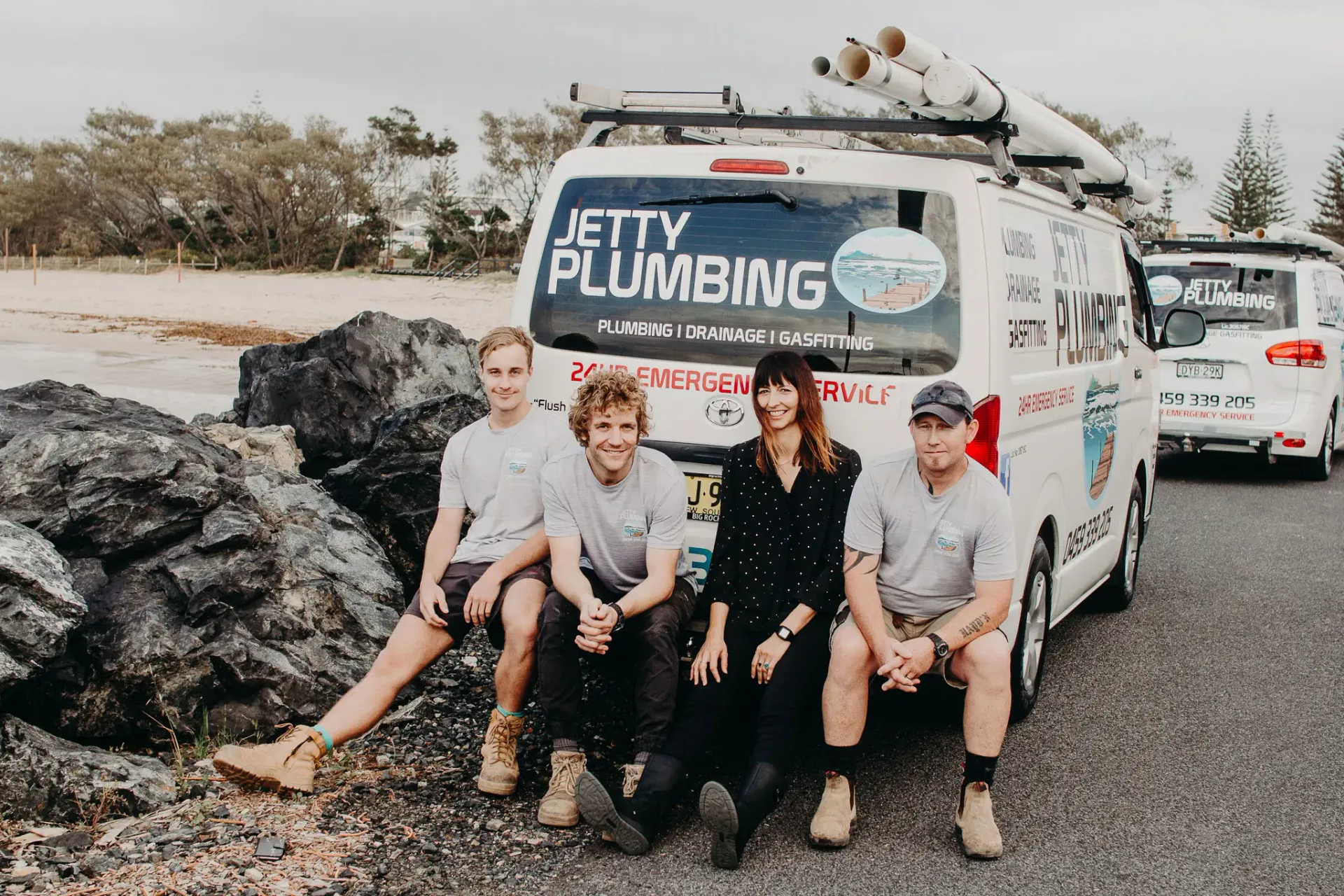 Four people in work clothes sit in front of a white Jetty Plumbing van parked by rocky terrain near a beach.