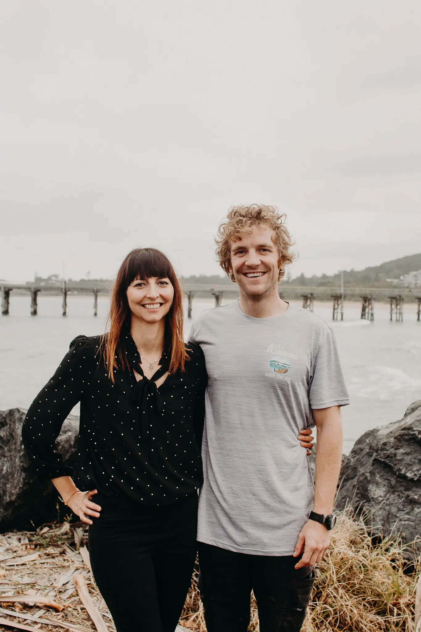A smiling couple standing on a rocky beach with a pier in the background under a cloudy sky.