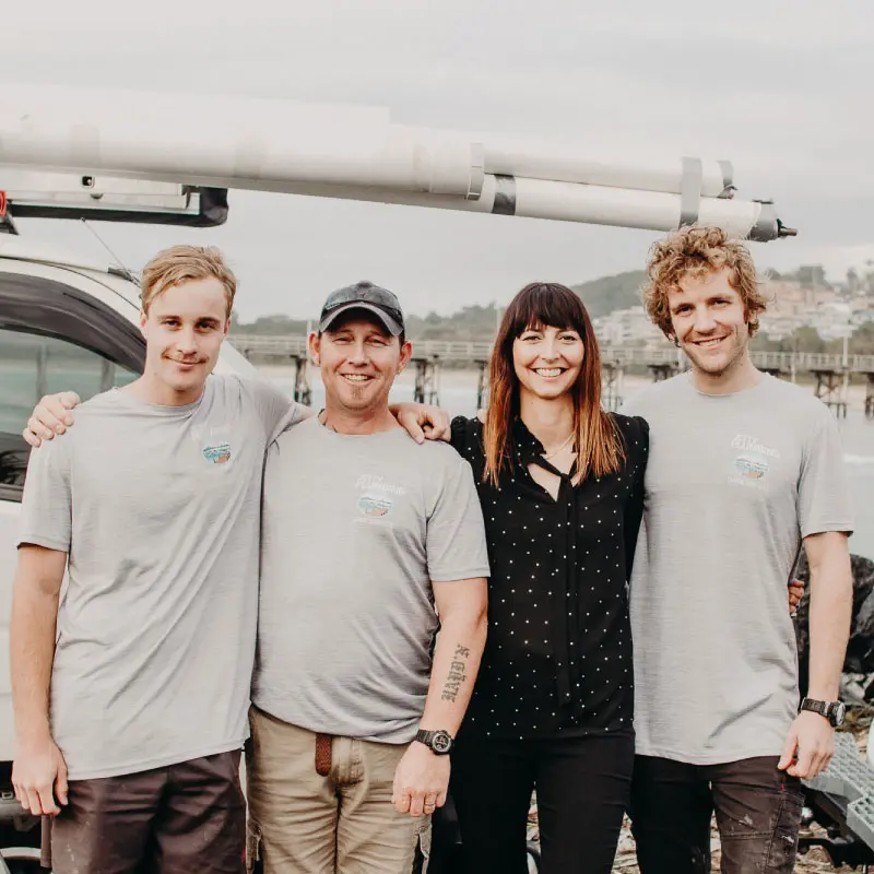 Four people smiling together outdoors in front of a vehicle with a roof rack, wearing casual attire.