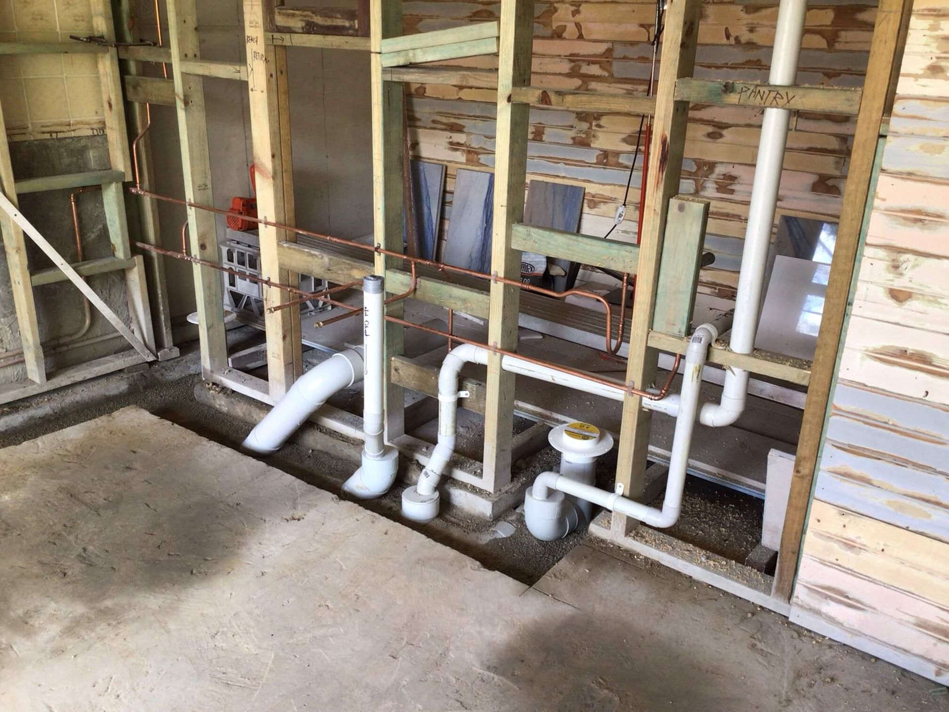Interior construction site showing white PVC plumbing pipes installed within exposed wooden wall studs on concrete floor.
