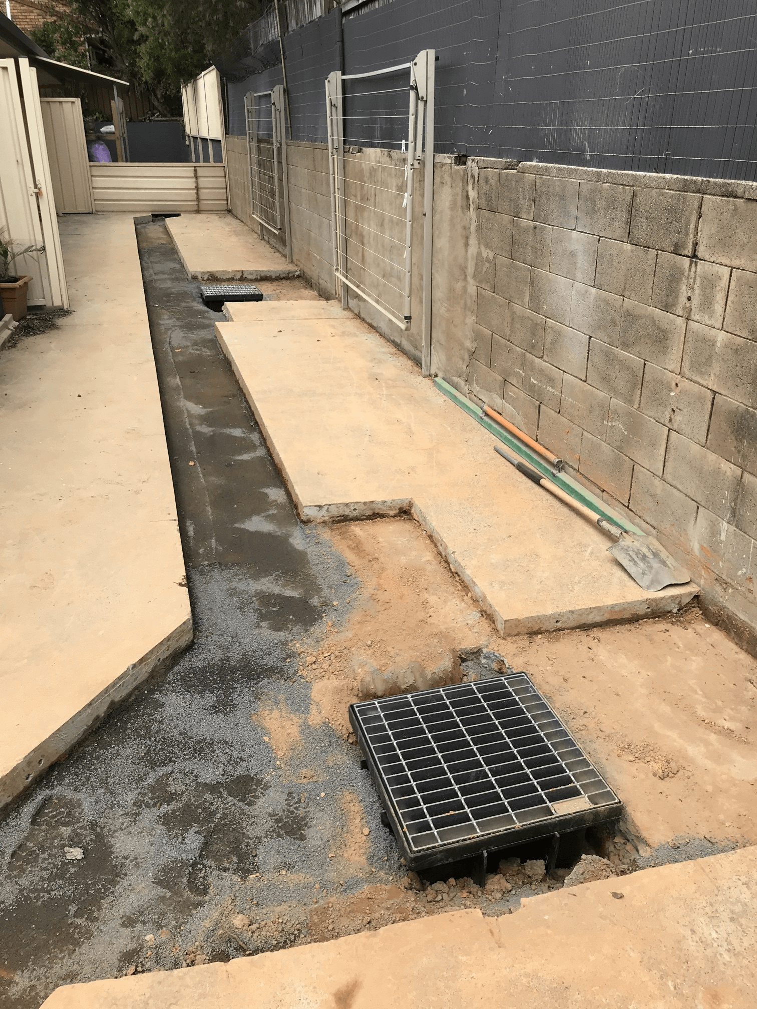 A narrow construction site between a concrete wall and a walkway with a rectangular storm drainage grate and gravel patch.