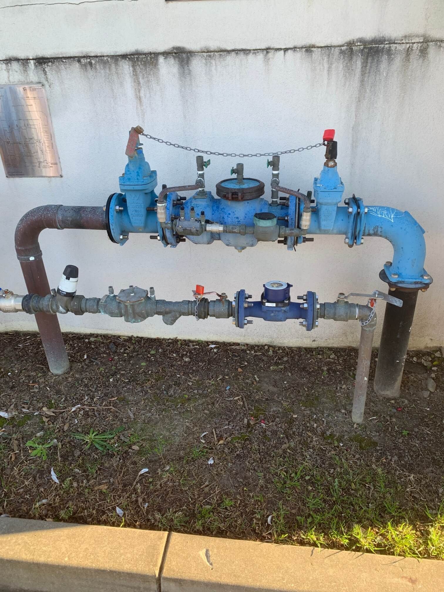 A water backflow prevention assembly with two parallel blue pipe systems against an outdoor white wall.