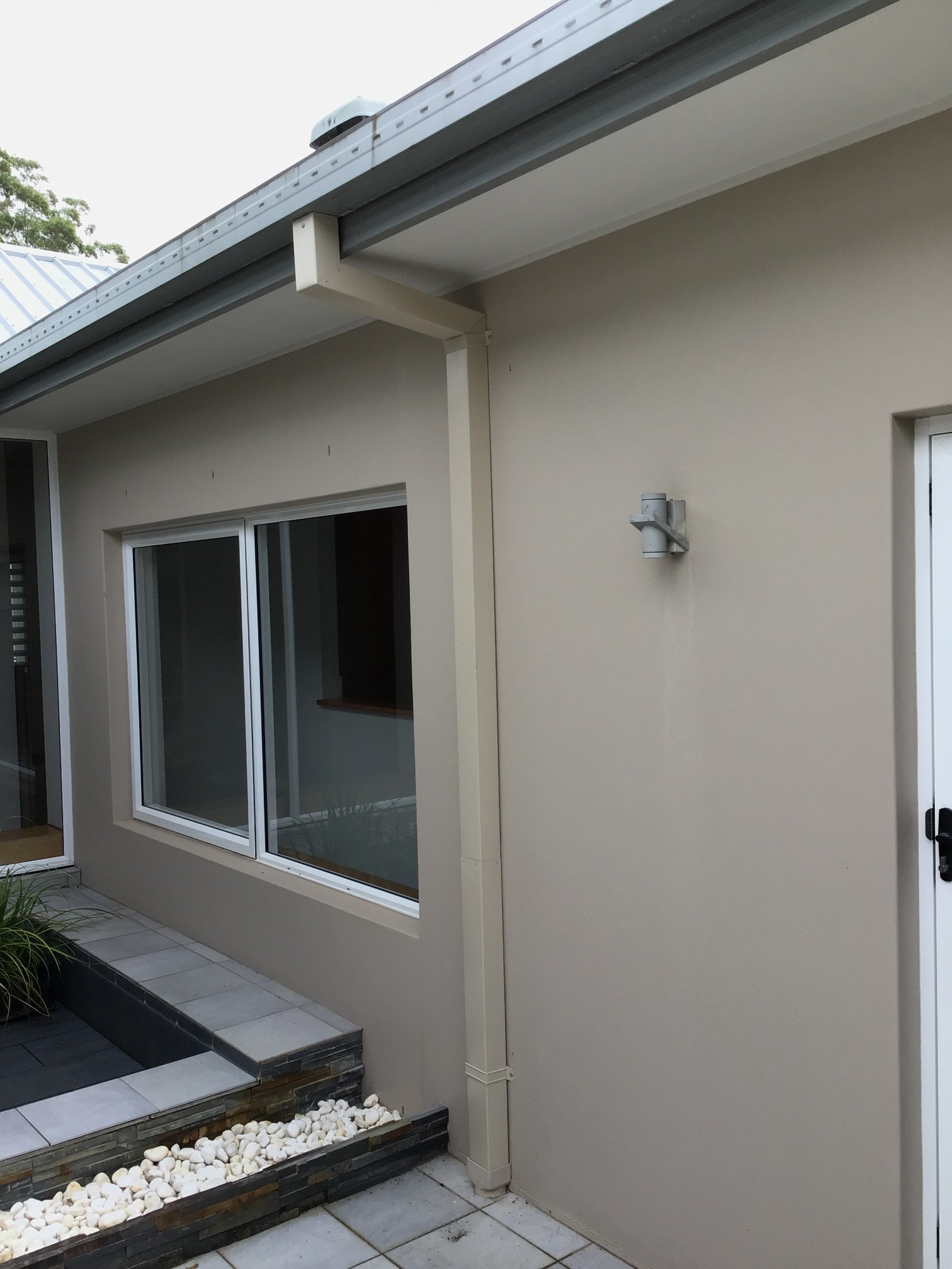 A beige exterior wall of a house with a vertical downpipe and a rectangular window overlooking a small stone-filled patio.