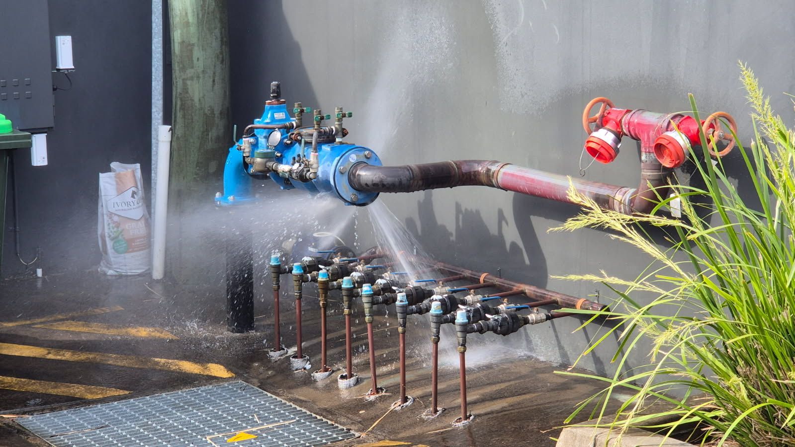 A broken pipe spraying water near a wall, featuring blue valves and a red pipe connection.