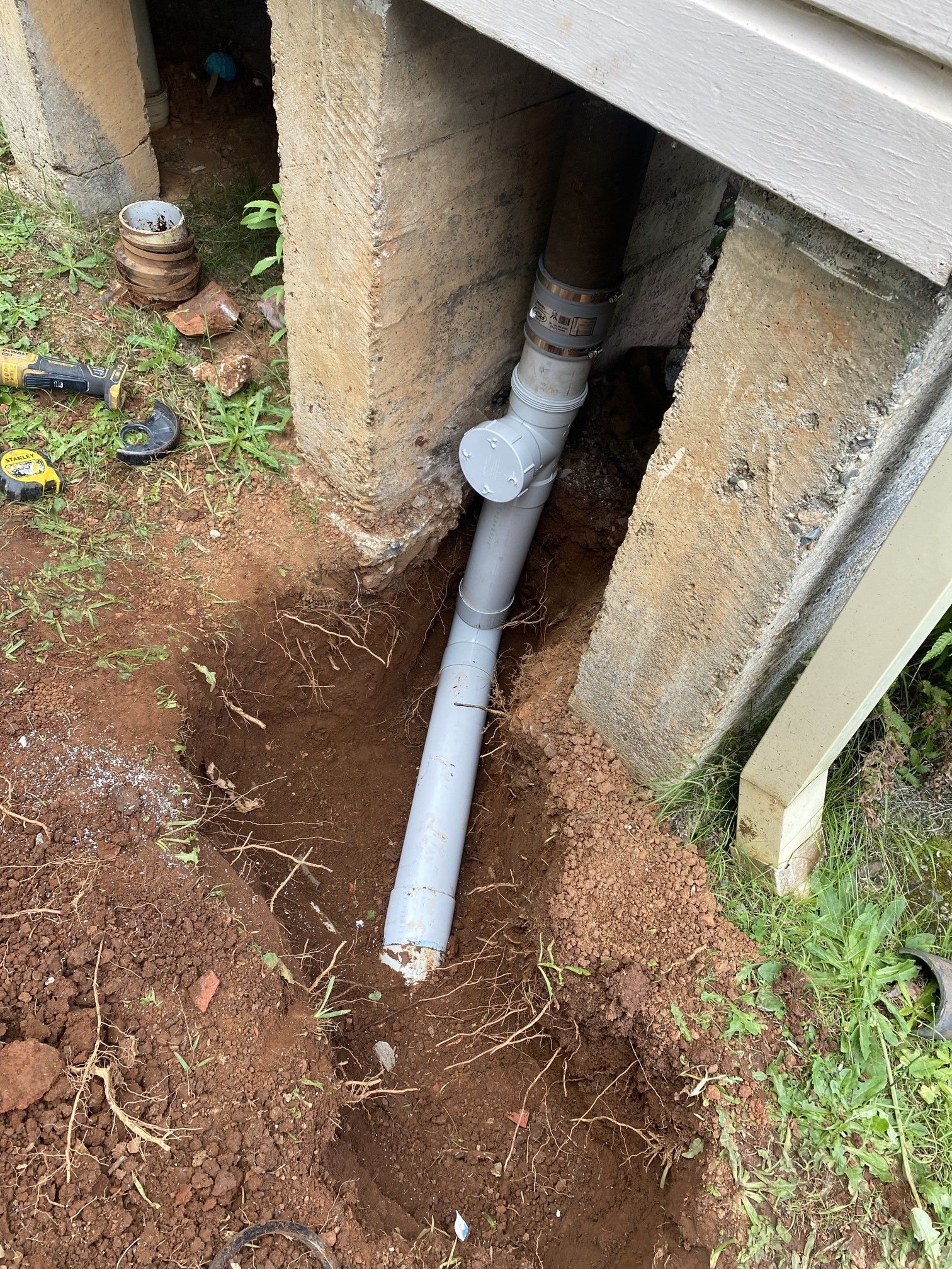 A gray plastic drain pipe extension connected to a vertical pipe under a house foundation, set in a trench of dirt.