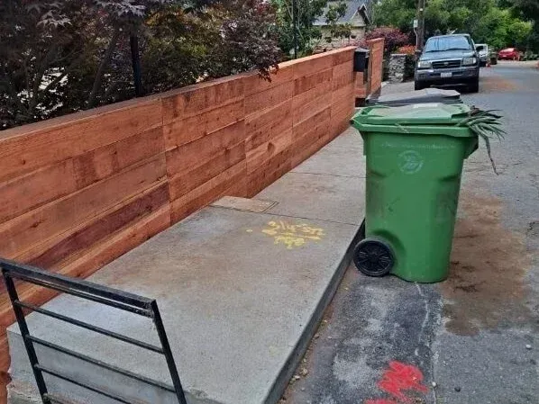 Sidewalk with a green trash bin, low brick wall, and parked cars along a street.