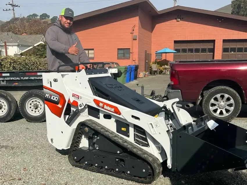 Man standing on a white tracked skid steer near a red pickup in a gravel lot by a brick building