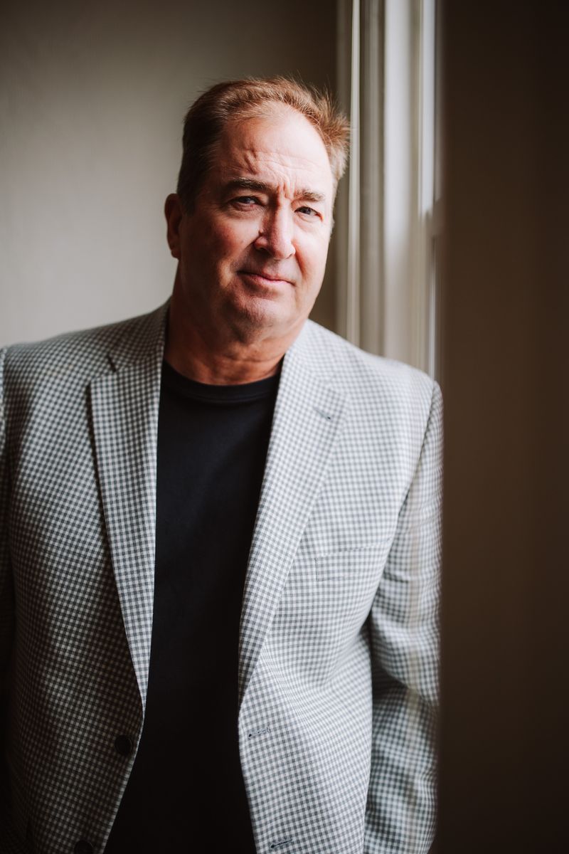 Man in a light patterned blazer standing by a window, looking at the camera.