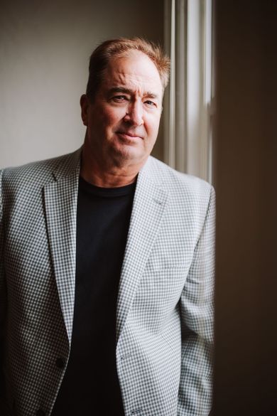 Man in a light patterned blazer standing by a window, looking at the camera.