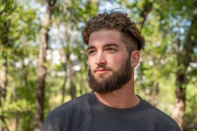 Bearded man in a dark shirt standing outdoors with trees blurred in the background