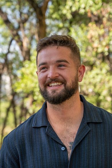 Smiling man in a dark blue shirt outdoors with blurred trees in the background