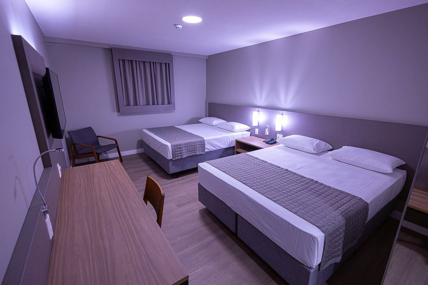 Quarto de hotel com duas camas de solteiro, escrivaninha e TV. Decoração em tons de marrom claro e bege.