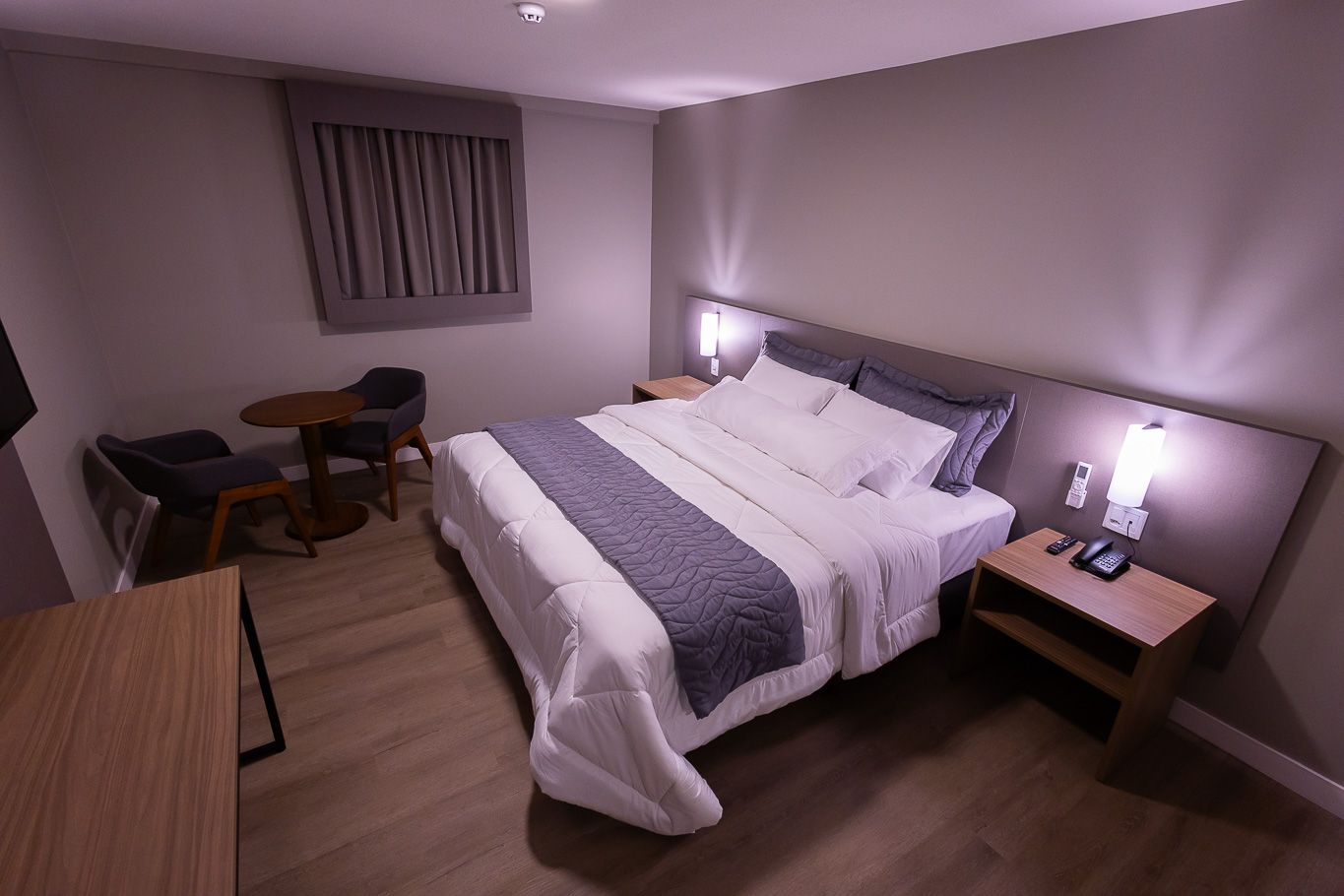 Quarto de hotel com cama, mesas de cabeceira, uma mesinha e cadeiras.