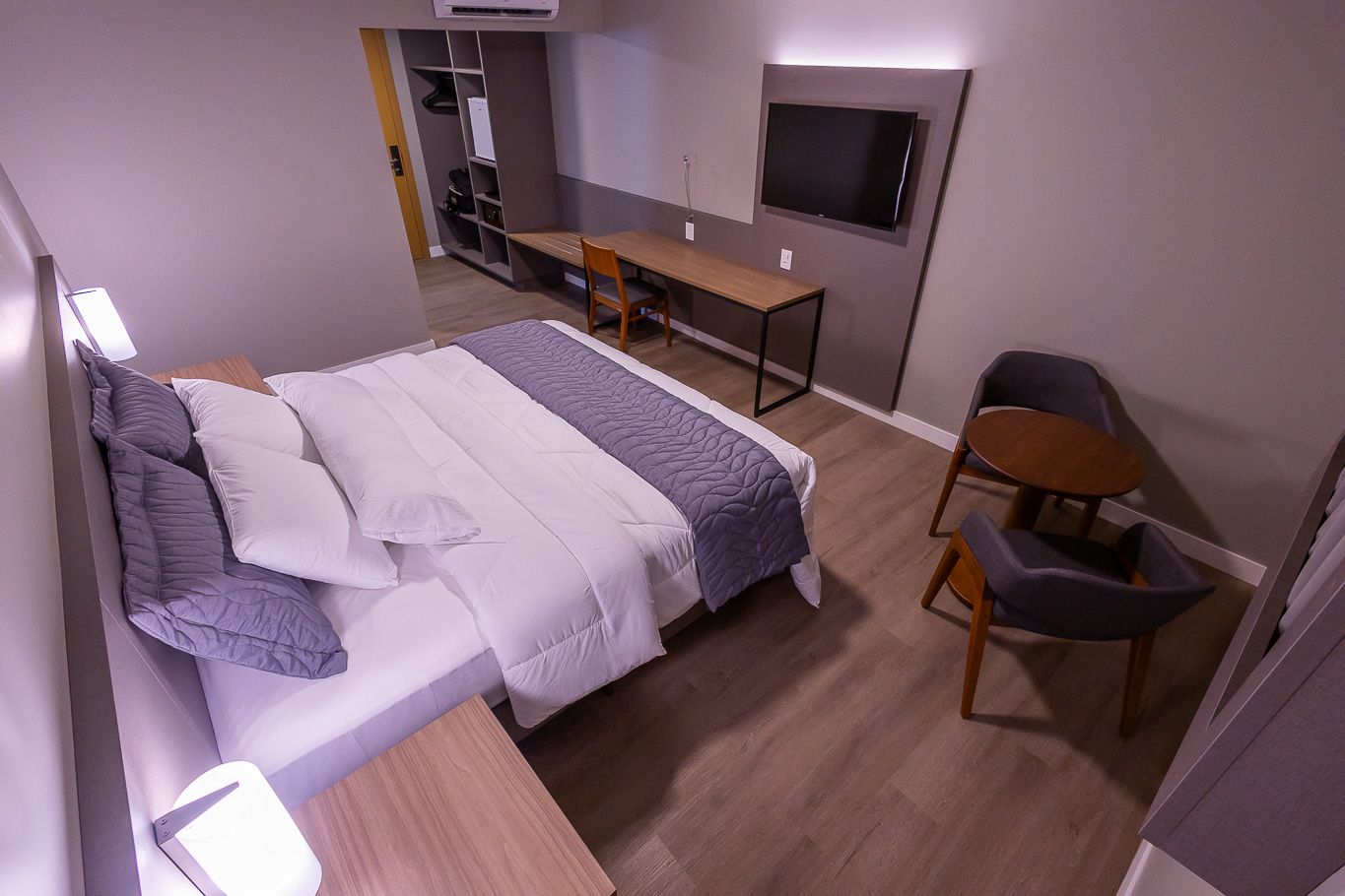 Quarto de hotel com cama, escrivaninha, cadeiras e televisão. Paredes cinza, piso de madeira e mobília moderna.
