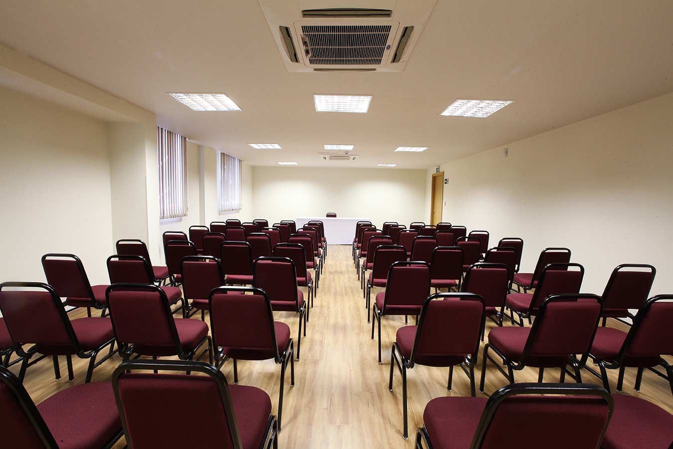 Uma sala de conferências com fileiras de cadeiras cor de vinho em frente a uma mesa sob luzes de teto.
