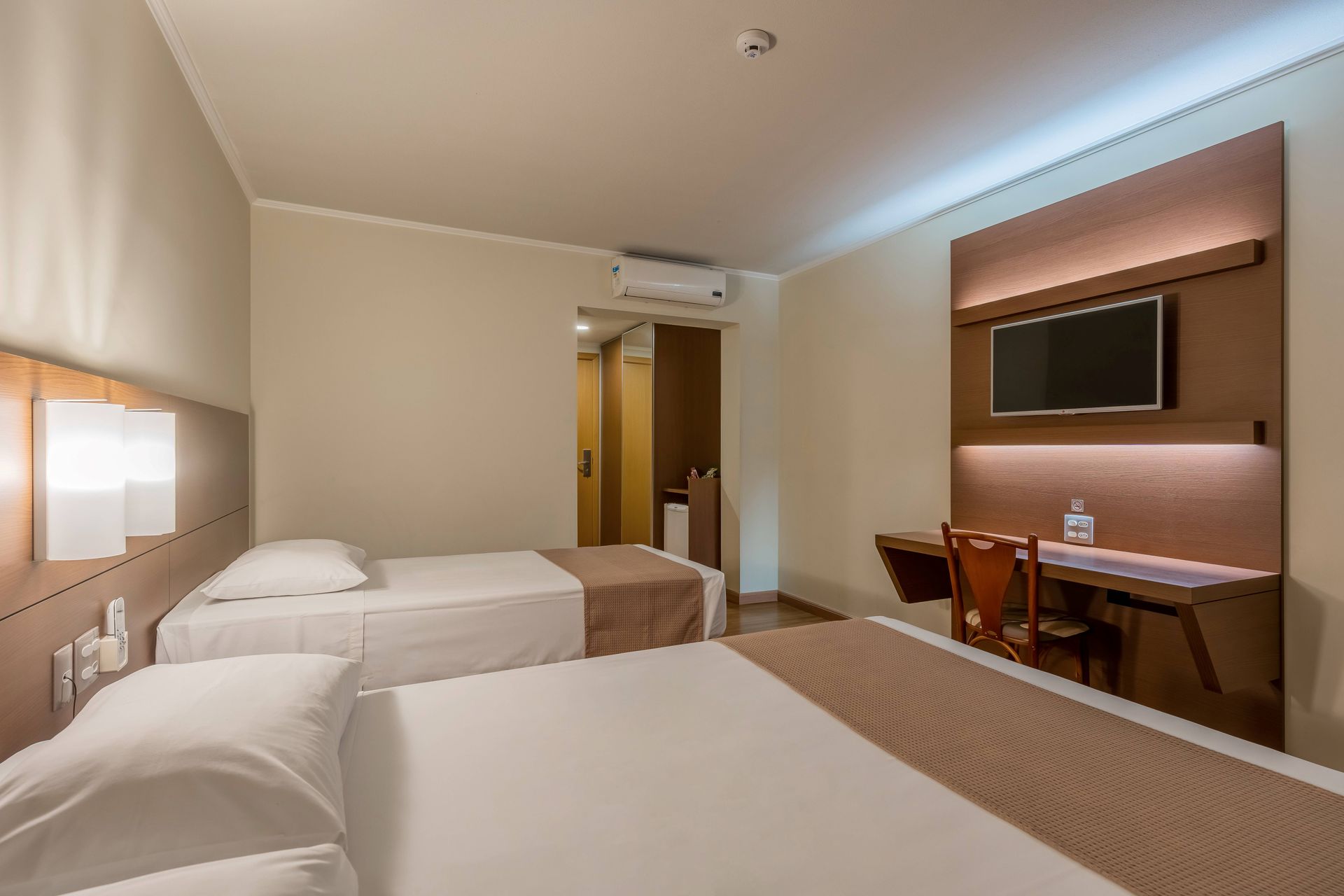 Quarto de hotel com duas camas, escrivaninha e TV na parede. Tons de creme, marrom e bege.
