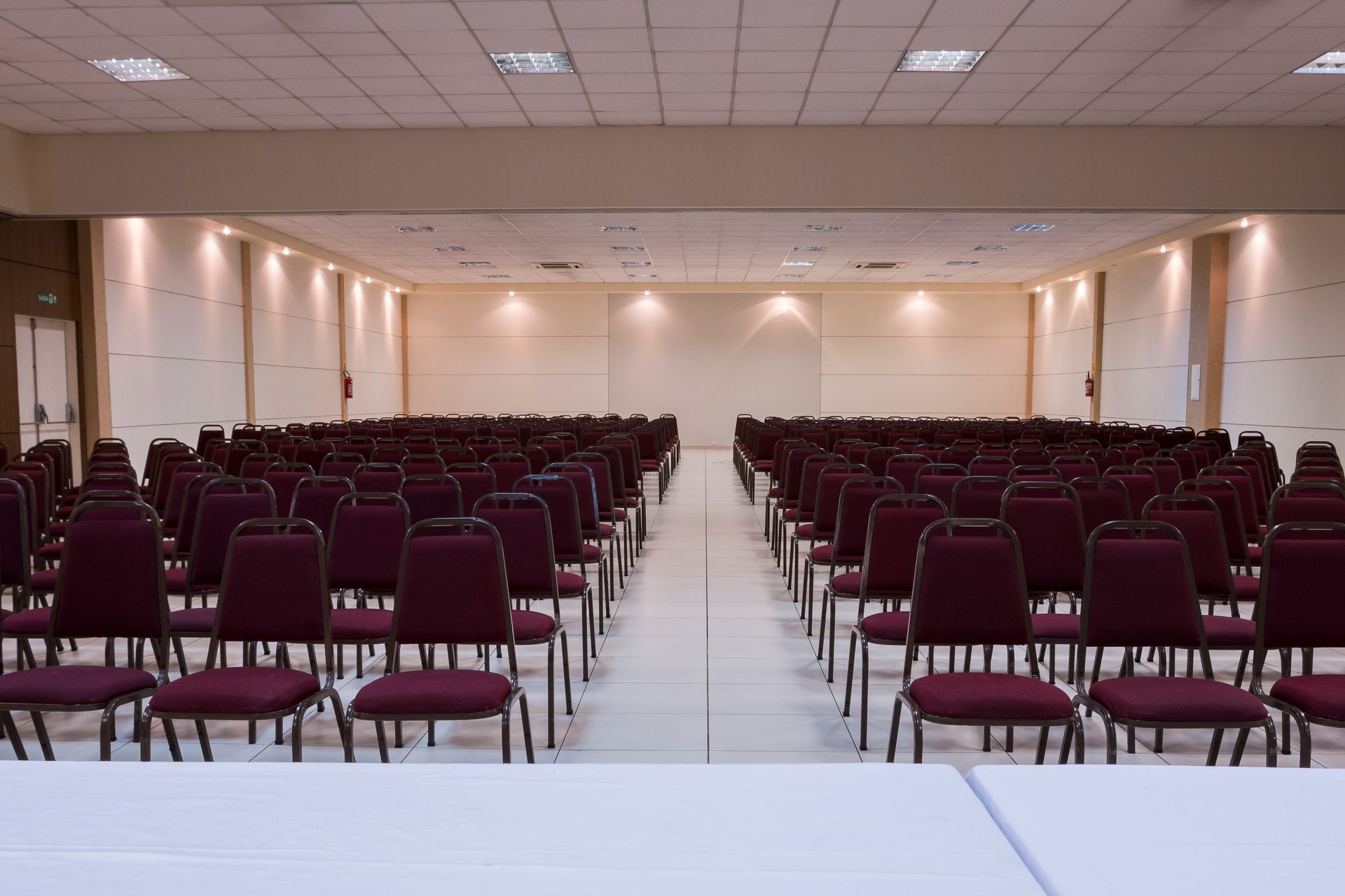 Fileiras de cadeiras vermelhas preenchem uma grande sala de conferências vazia, com uma parede branca e holofotes.
