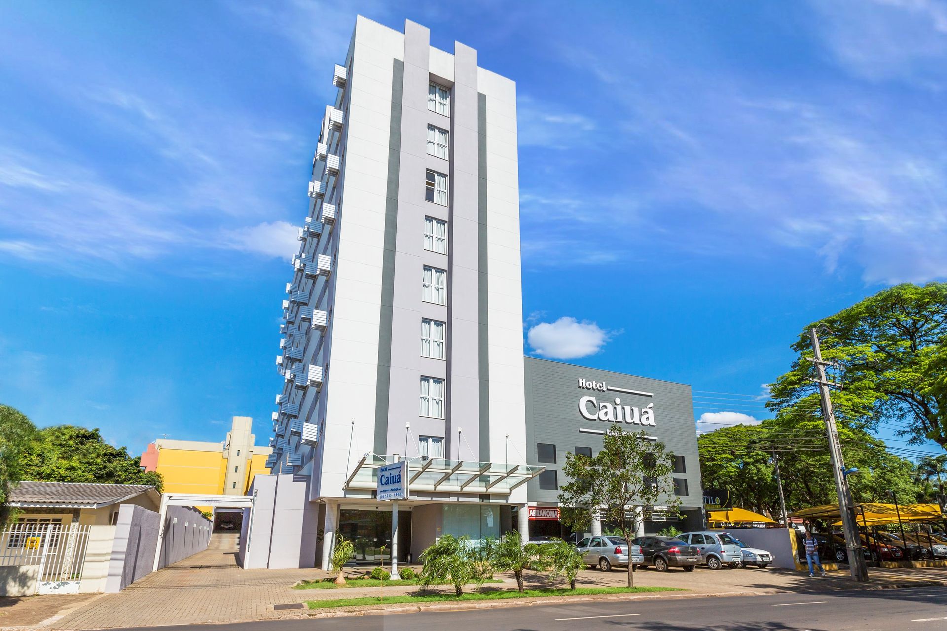 Hotel alto, de vários andares, com fachada branca e cinza, sob um céu azul. Placa com o nome 