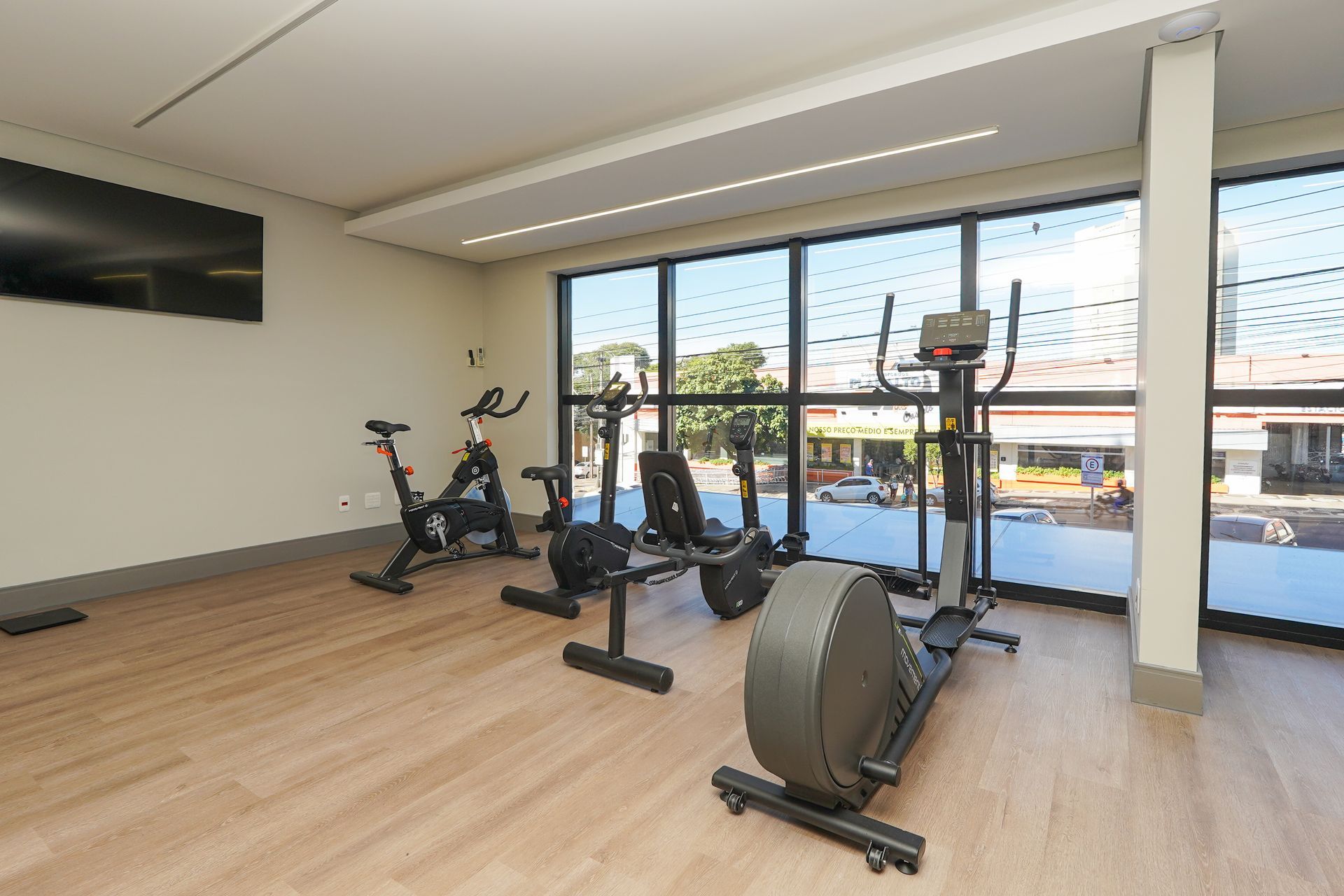 Sala de ginástica com bicicletas ergométricas, máquinas de remo e grandes janelas com vista para uma varanda.