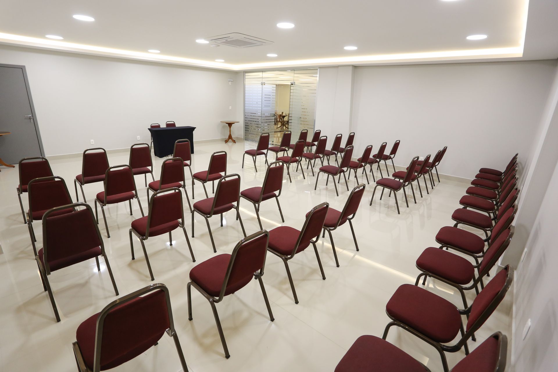 Sala de conferências vazia com fileiras de mesas e cadeiras pretas, paredes e teto brancos.