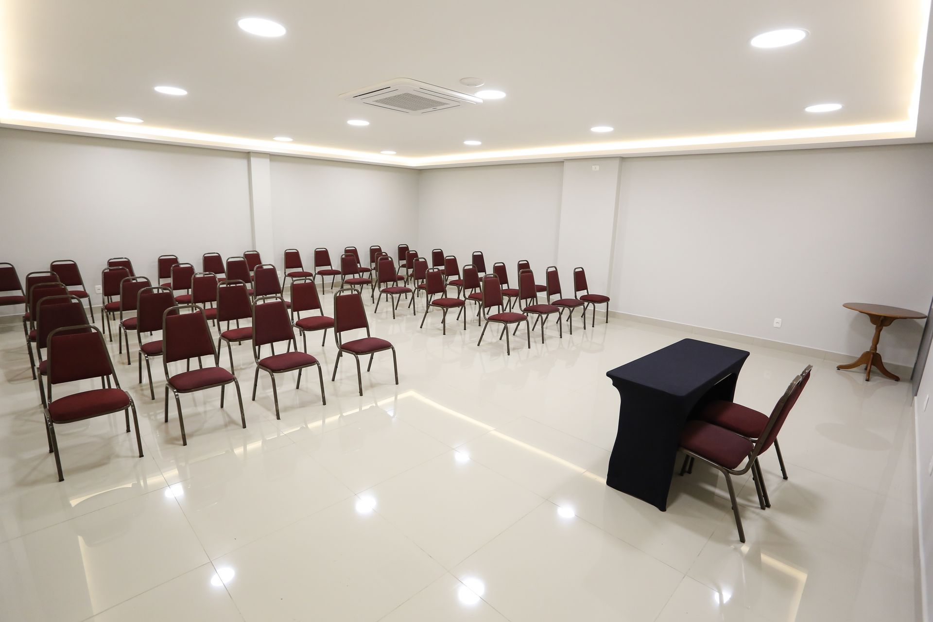 Sala de conferências com mesas em formato de U, toalhas de mesa azul-escuras, cadeiras cinza e iluminação embutida.