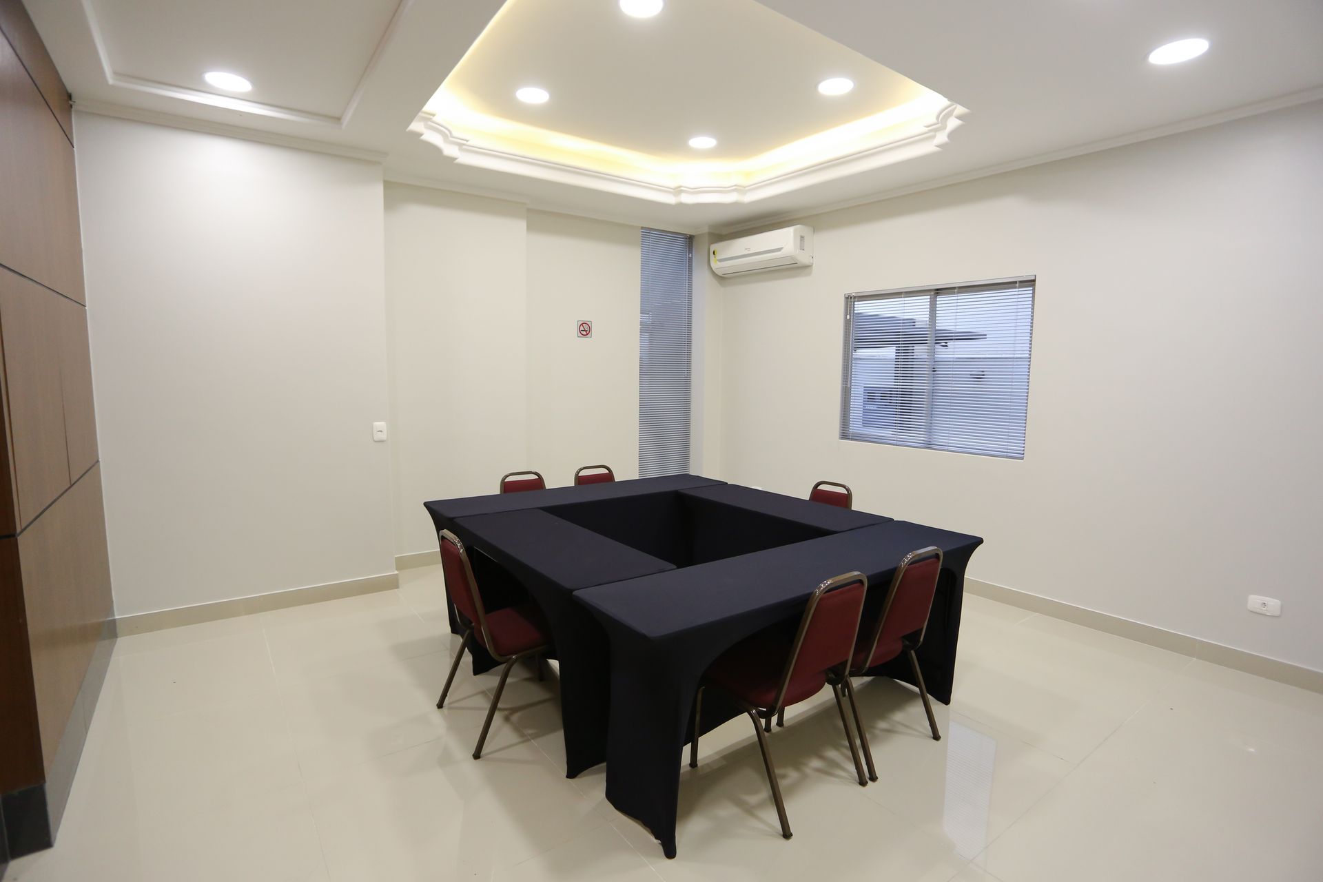 Sala de conferências com mesa e cadeiras em formato de U. Paredes e teto em tons claros.