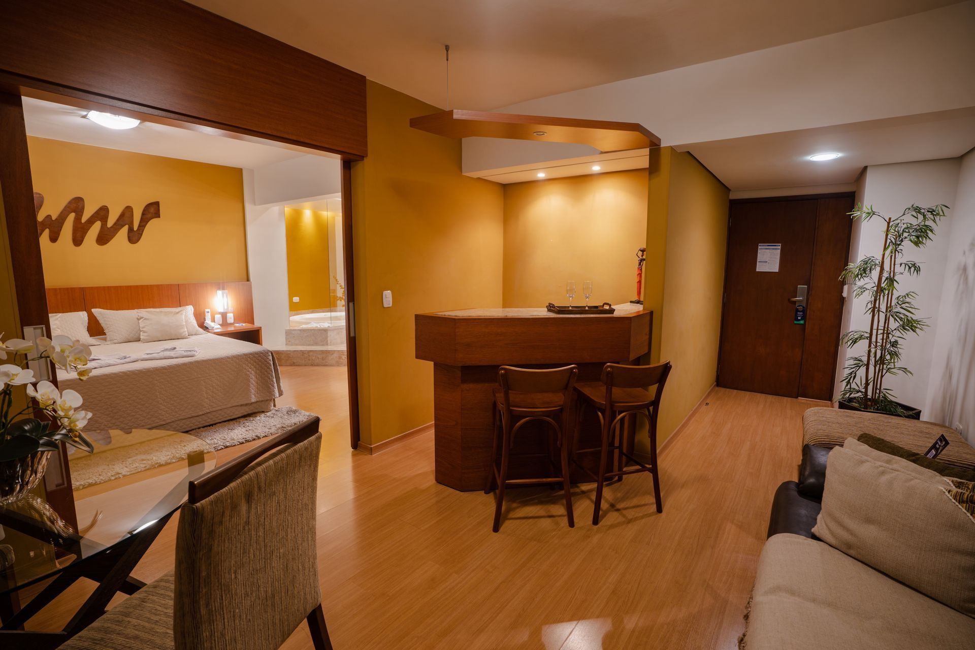 Quarto de hotel com bar, quarto e sala de estar em tons quentes.