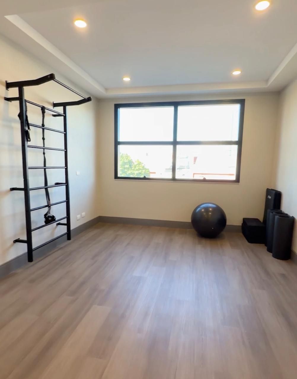 Quarto de hotel com cama, escrivaninha com TV, cadeira e sofá preto. Cortinas bege e piso de madeira.