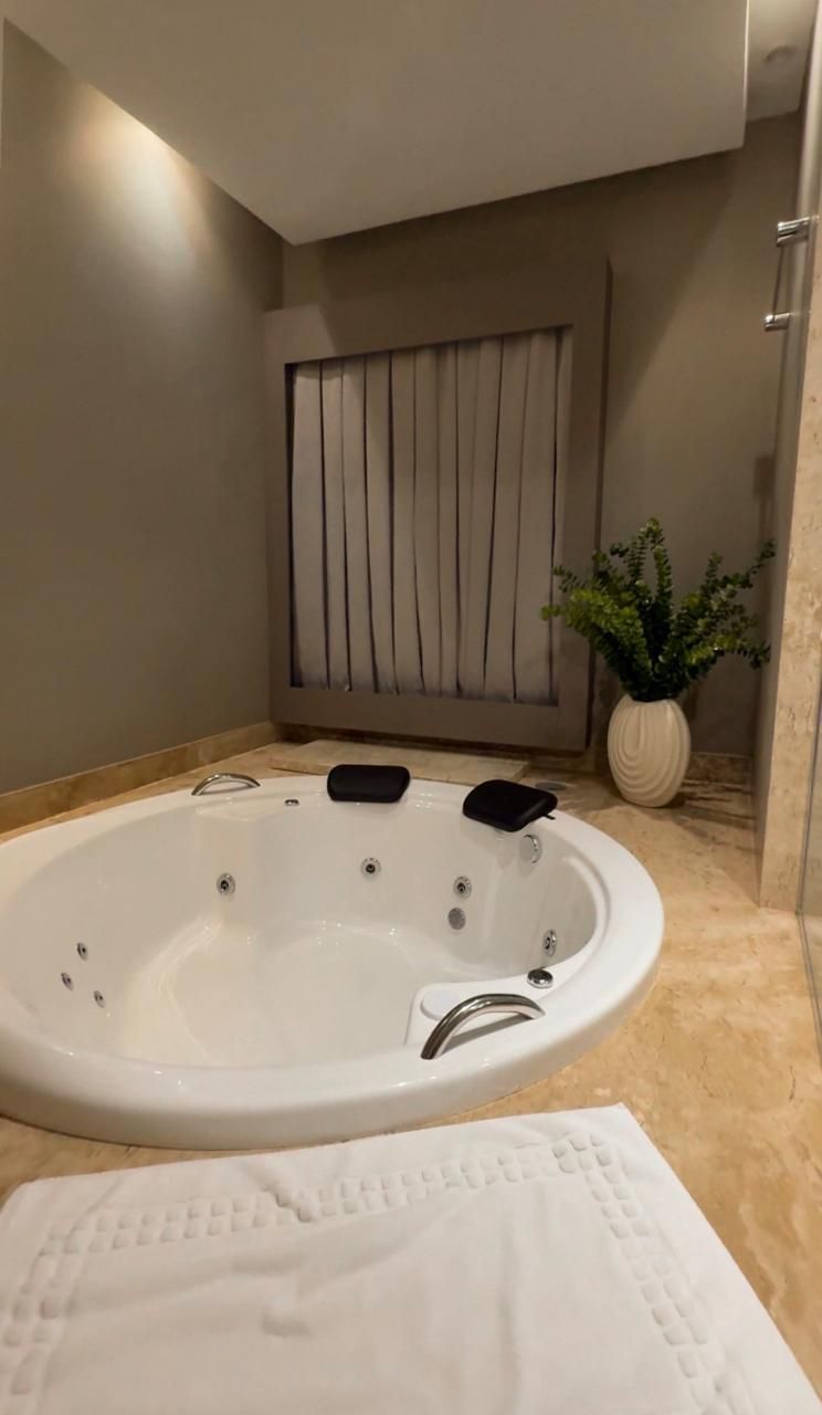Quarto de hotel com três camas, uma escrivaninha e uma porta. Piso de madeira clara, paredes em tons neutros e peseiras bege.