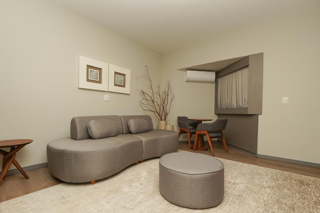 Suíte nupcial com Sala de estar aconchegante em tons de bege com sofá curvo, pufe redondo, escrivaninha pequena e obras de arte na parede sob iluminação quente.