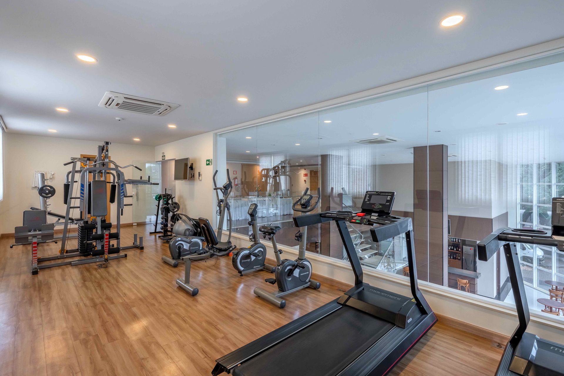 Academia com diversos aparelhos de ginástica, piso de madeira e parede de vidro que exibe equipamentos adicionais.