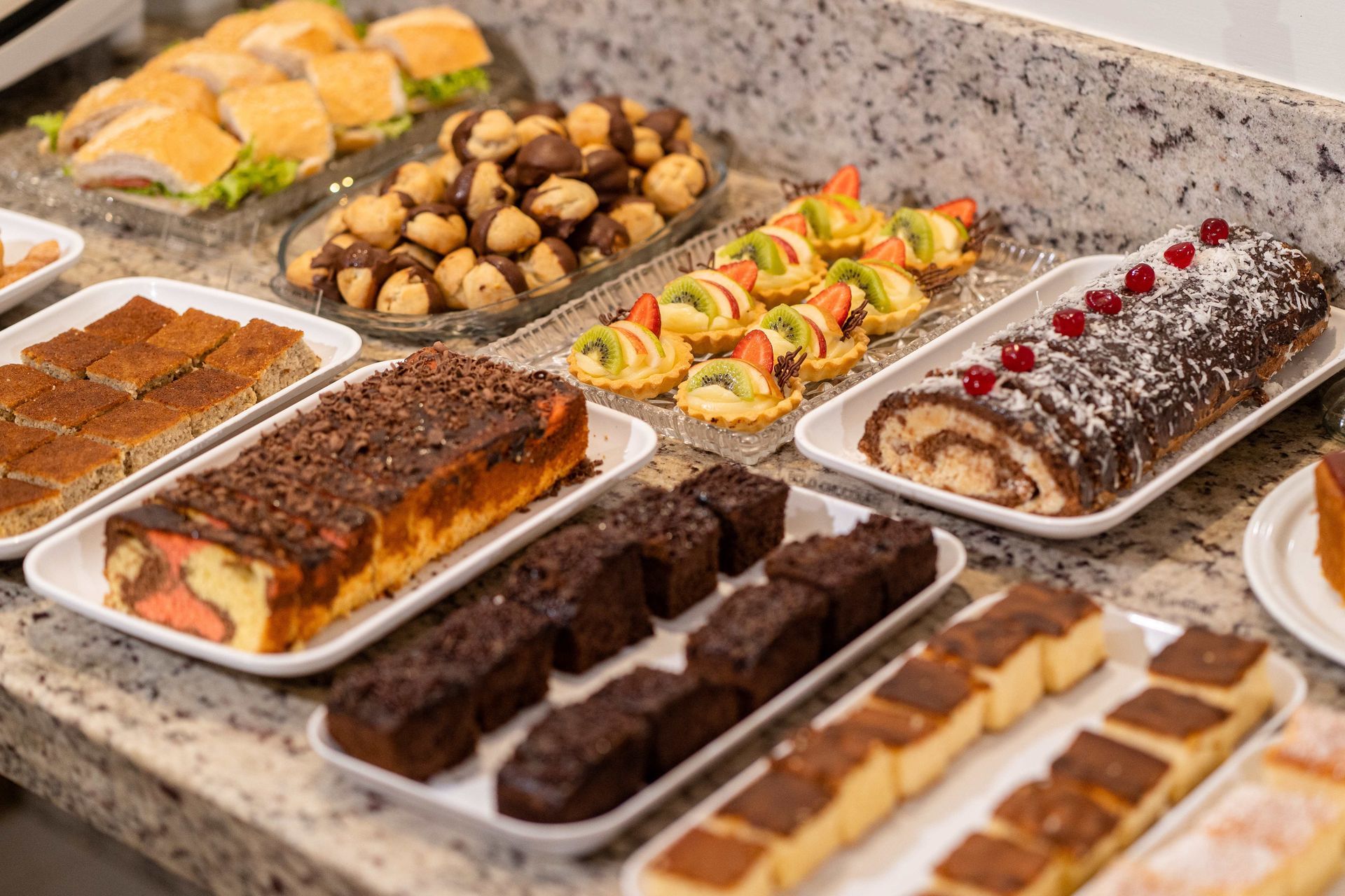 Exposição de sobremesas com sanduíches, biscoitos, doces, tortas de frutas, brownies e um rocambole em uma bancada.